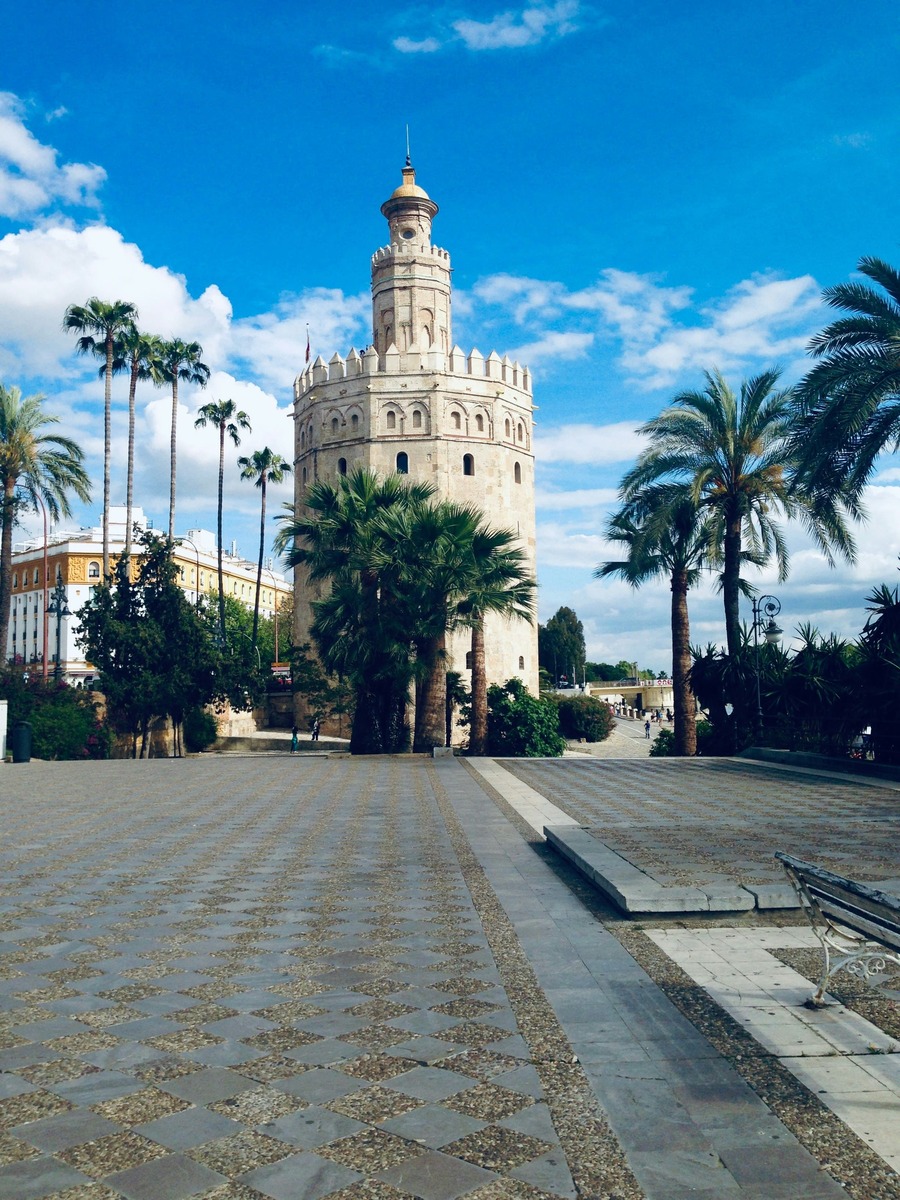 La Torre del Oro vista desde una plaza empedrada con palmeras bajo un cielo azul con nubes